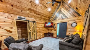 Acogedor interior de una cabaña de troncos con chimenea, dos sillones de cuero, un sofá, un televisor y un reloj en la pared.