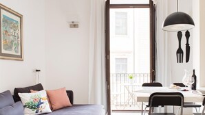 A modern living room with a grey sofa, a dining table with chairs, a balcony with a railing, and a painting on the wall. - Suite La Piazza by Wonderful Italy (Syracuse)