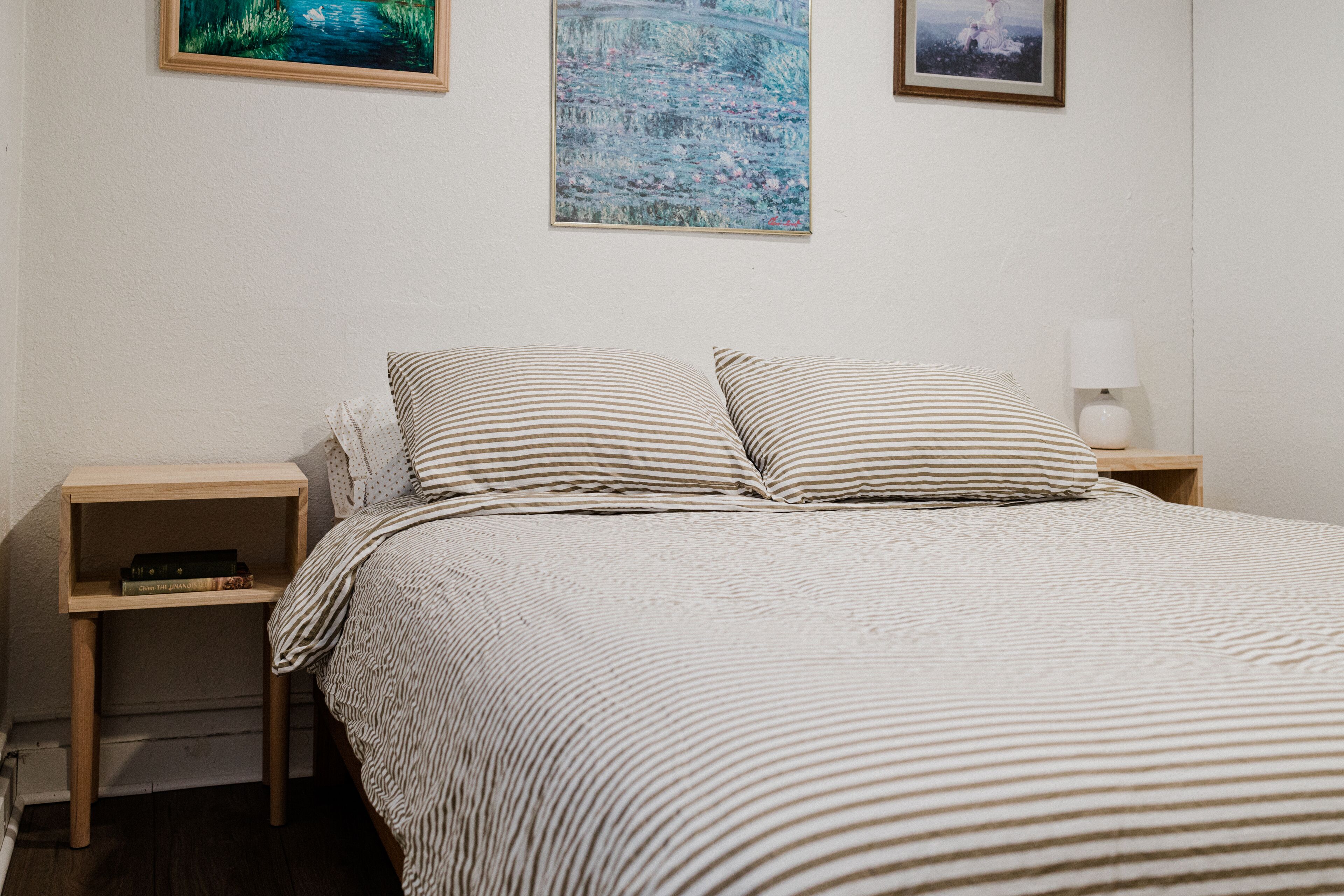 Ein ordentlich bezogenes Bett mit gestreifter Bettwäsche, zwei Nachttische mit Büchern und Wandbilder über dem Bett.