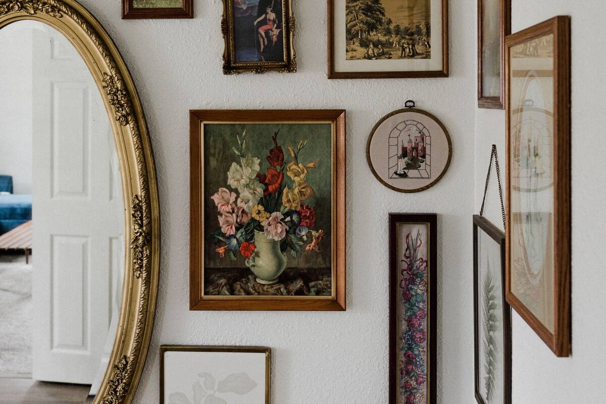 Una habitación con un espejo grande, varias obras de arte enmarcadas, una silla de mimbre y un sillón blanco mullido.