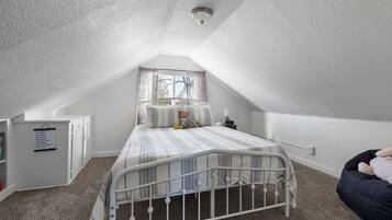 Um quarto com teto inclinado, uma cama com colcha listrada, um urso de pelúcia, uma mesa de cabeceira e uma janela com cortinas.
