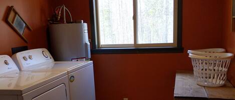 Un lavadero con lavadora y secadora, una ventana que da a árboles, un letrero sobre la ventana y una pequeña mesa con una cesta.