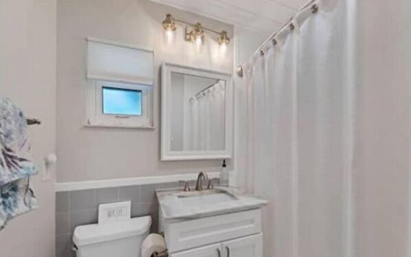 A bathroom with a toilet, sink, and mirror. There is a window above the toilet and a shower curtain.