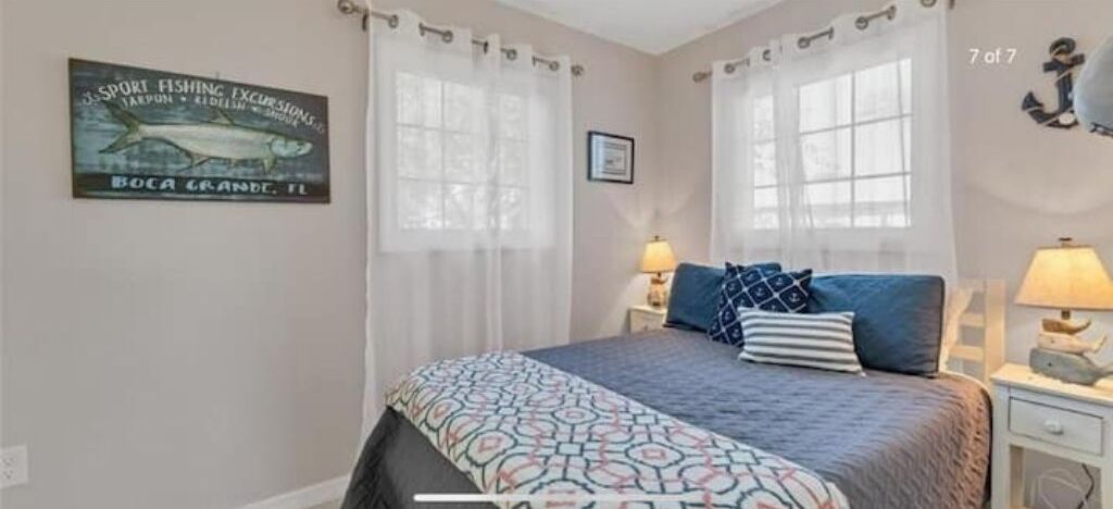 Ein Schlafzimmer mit einem Bett, zwei Nachttischen mit Lampen, einem Fenster mit Vorhängen, einem gerahmten Bild an der Wand und einem Schild an der Wand.