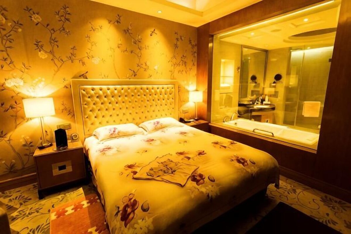 A hotel room with a large bed, bedside lamps, a nightstand, a mirror, and a window with a view of the bathroom.