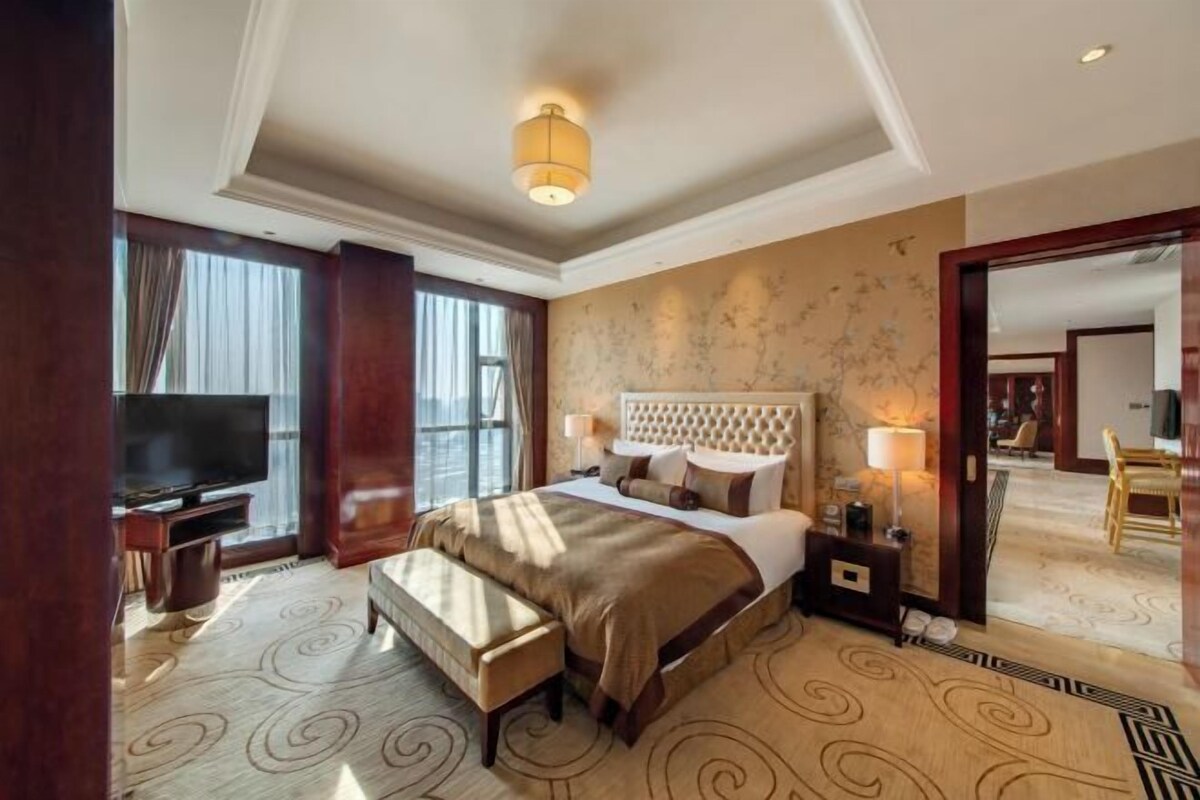 A hotel room with a large bed, a TV, a bench, and a view of the lobby.