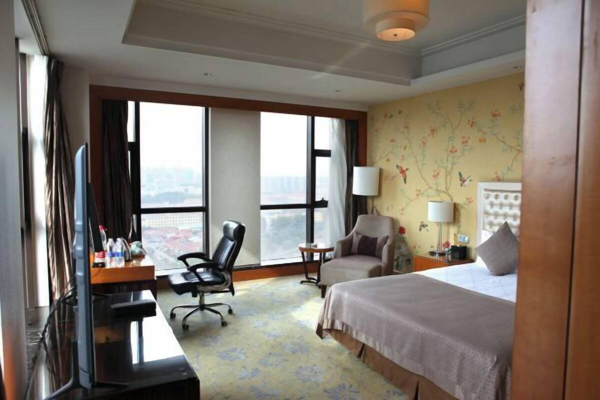 A hotel room with a large bed, two chairs, a desk, a TV, and a view of the city through large windows.
