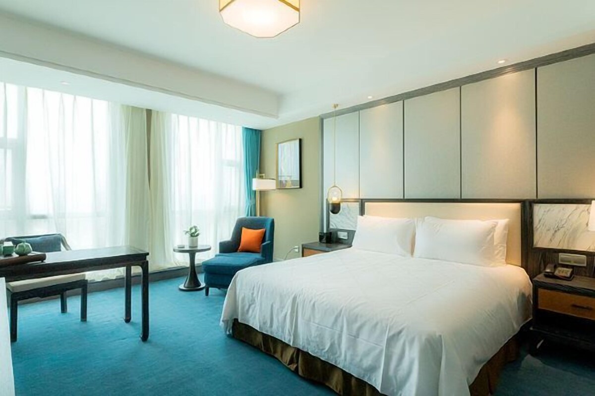 A hotel room with a large bed, a desk, a chair, a small table, and a window with curtains.