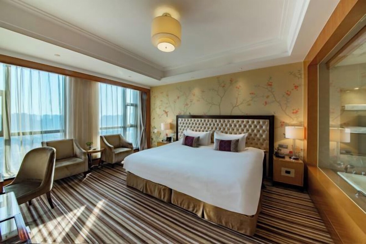 A hotel room with a large bed, a sitting area with a chair and a table, a nightstand with a lamp, and a view through the window.