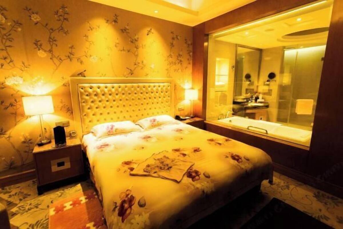 A hotel room with a large bed, a nightstand with a lamp, a mirror, and a bathroom visible through a glass partition.