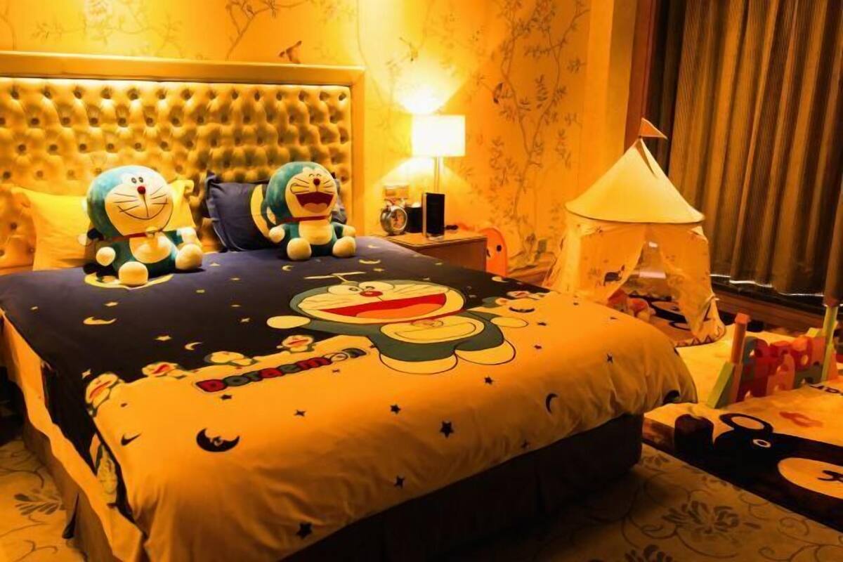 A hotel room with a tufted headboard, plush toys, a bed with a cartoon-themed bedspread, a bedside lamp, and a window with curtains.