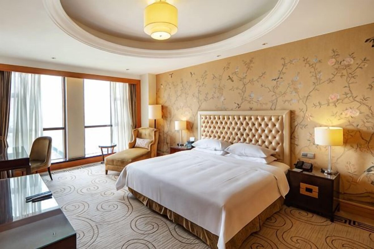 A hotel room with a large bed, a desk, and a chair. There are two lamps on the nightstands and a large window with curtains.
