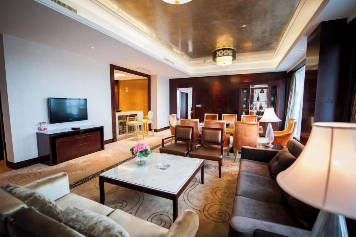 A hotel living room with a flat-screen TV, sofas, a coffee table, and a dining area with chairs and a table.