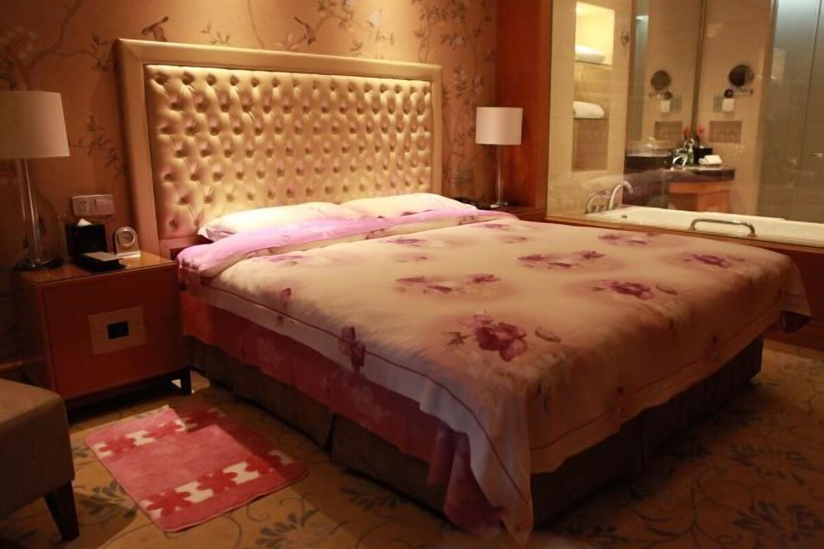 A bedroom with a tufted headboard, a bed with floral bedding, a nightstand with a lamp, a bathroom with a mirror, and a shower area.