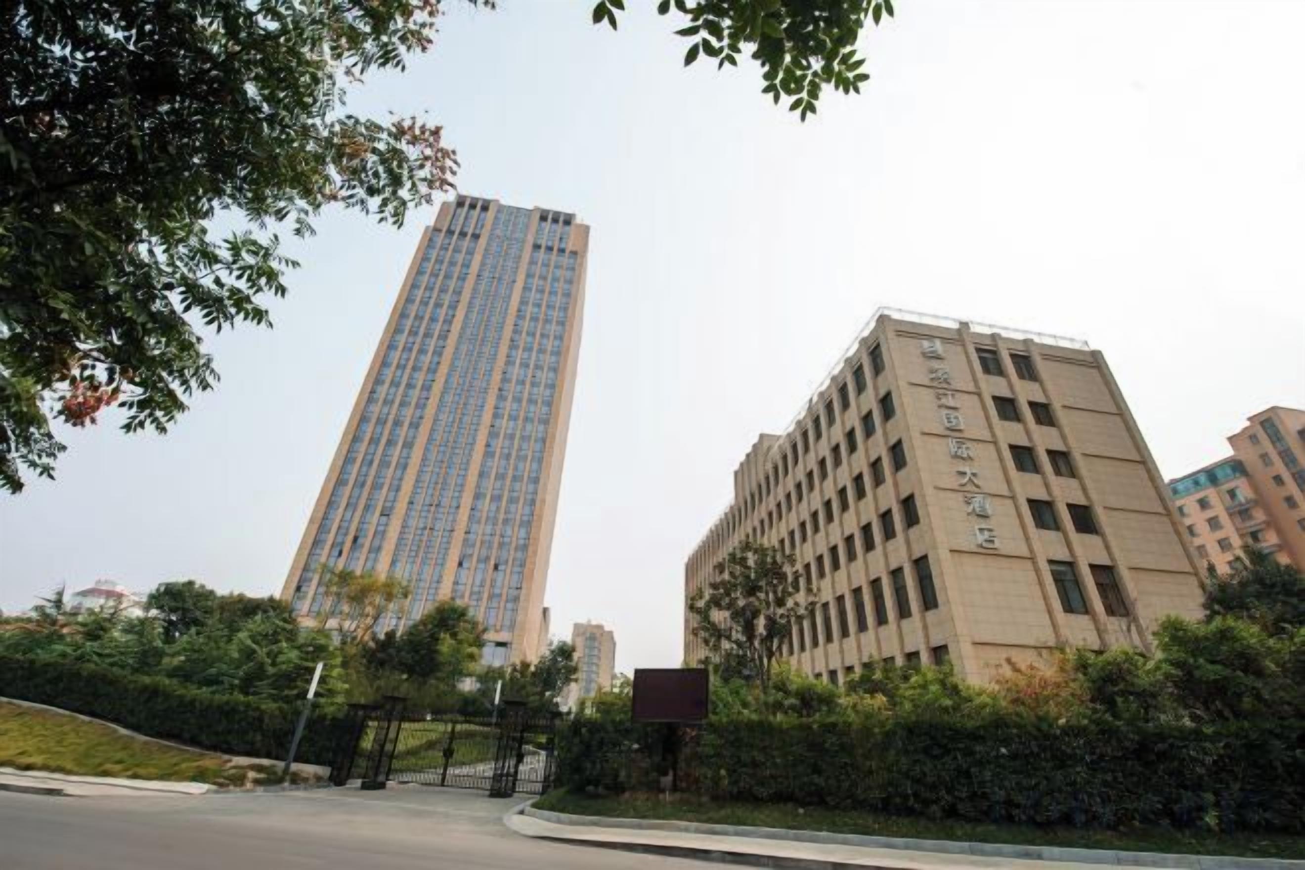 A tall building with a modern design, surrounded by greenery and other buildings.