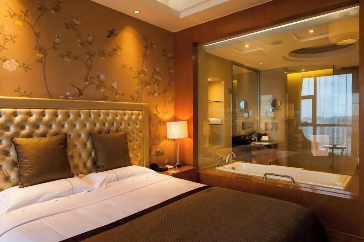 A hotel room with a tufted headboard, a large bed with white linens, a bedside lamp, a glass-enclosed bathroom with a bathtub, and a floral wall mural.