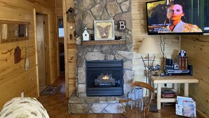 Ein gemütliches Blockhaus-Interieur mit Kamin aus Stein, Holzwänden, einem Fernseher und einem kleinen Tisch mit Büchern und Dekoration.