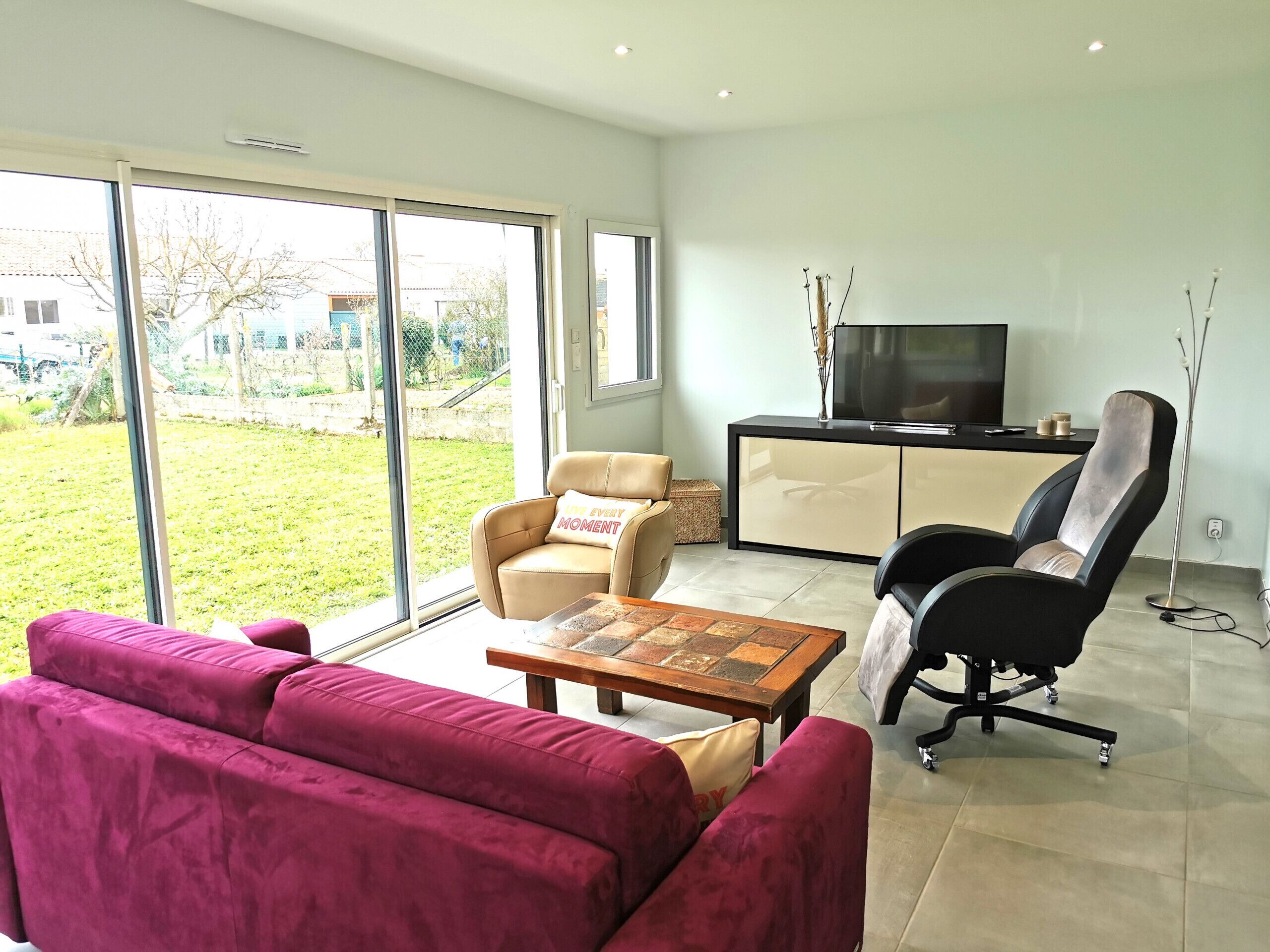 A modern living room with a large sliding glass door leading to a garden, a beige sofa, a red sofa, a black office chair, and a wooden coffee table.
