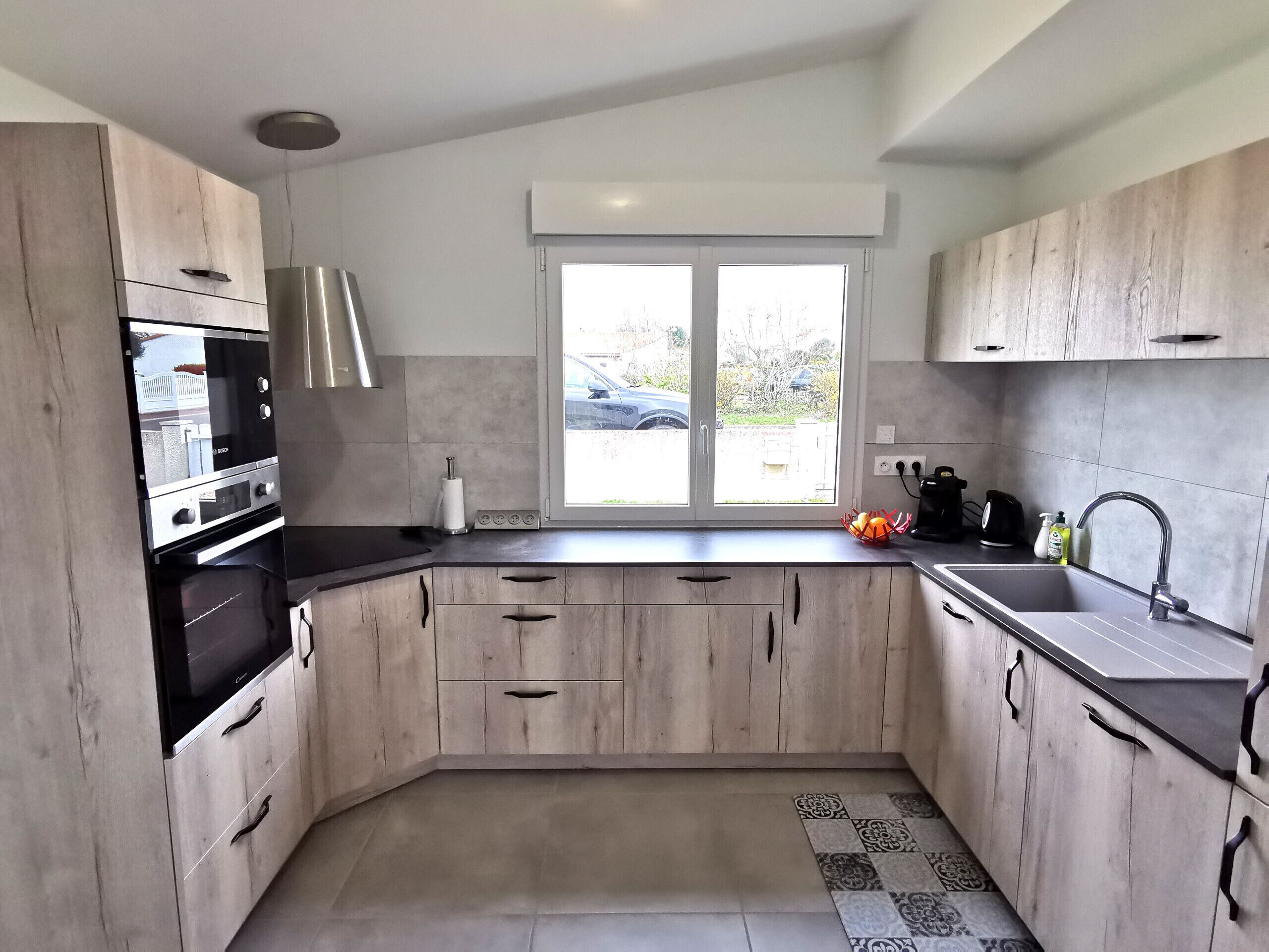 A modern kitchen with light wood cabinets, a built-in oven, and a large window.