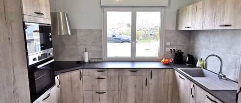 A modern kitchen with light wood cabinets, a built-in oven, and a large window.