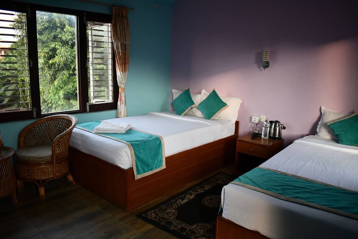 A room with two beds, a wicker chair, a nightstand with a kettle, and a view of greenery through the window.