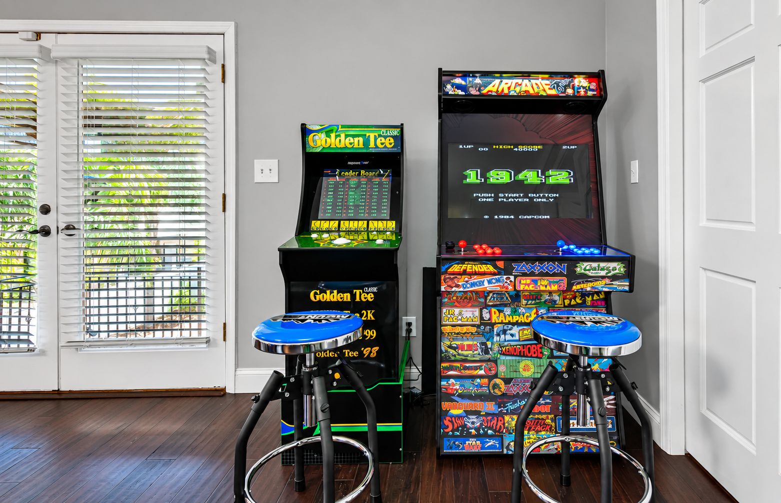 A room with two arcade machines, a door with blinds, and a wooden floor.