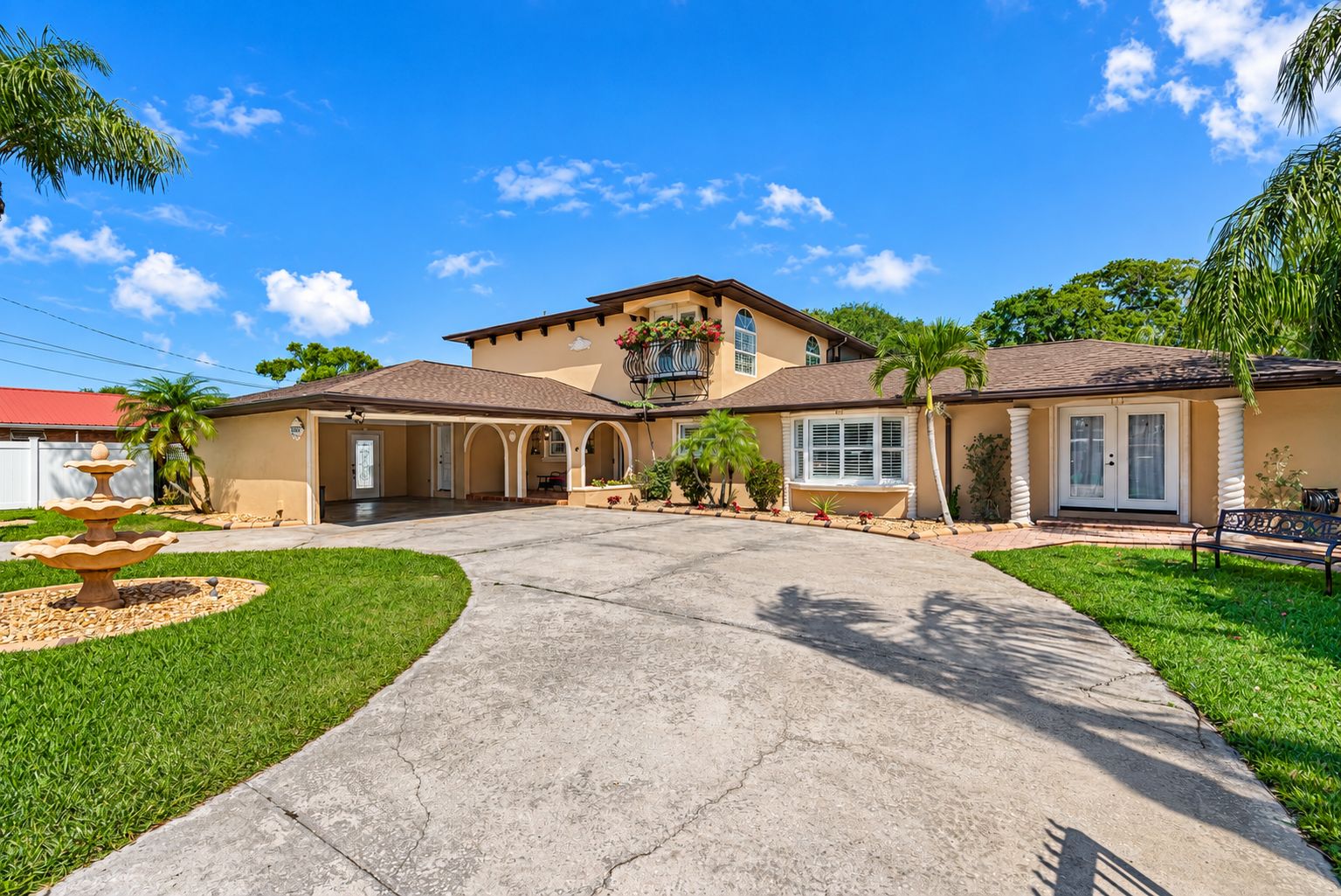 A two-story house with a driveway leading to a gated entrance, surrounded by well-maintained lawns and palm trees.