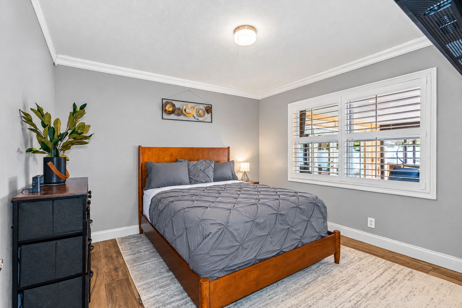 A bedroom with a bed, a nightstand, a potted plant, a wall-mounted artwork, and a window with blinds.