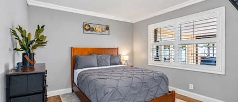 A bedroom with a bed, a nightstand, a potted plant, a wall-mounted artwork, and a window with blinds.