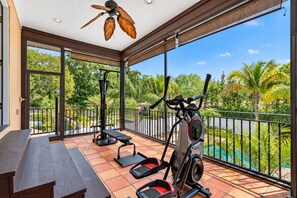 Ein Balkon mit Trainingsgeräten, einem Deckenventilator und Blick auf ein Schwimmbecken und Grünflächen.