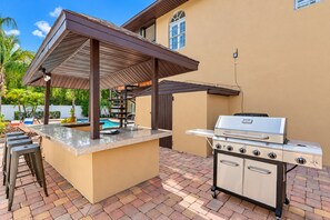 Un área de cocina al aire libre con una parrilla, taburetes de bar y una piscina.