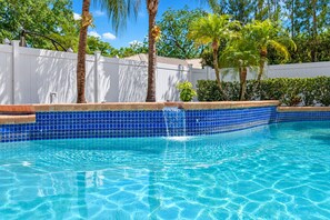 Una piscina azul cristalina con borde de azulejos, rodeada por una valla blanca y palmeras.