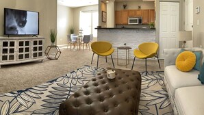 A living room with a television, a brown tufted ottoman, yellow chairs, a white sofa, and a patterned rug. - Cozy, bright 2BD w/yard, close to DT-bikes avail! (Manassas)