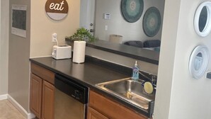 A compact kitchen with a black countertop, a built-in dishwasher, a sink with a soap dispenser, and a wall-mounted 'eat' sign. - Cozy, bright 2BD w/yard, close to DT-bikes avail! (Manassas)
