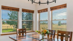 Ein Esszimmer mit Holztisch, vier Stühlen, einer Chandelüster und großen Fenstern mit Blick auf eine grüne Landschaft.