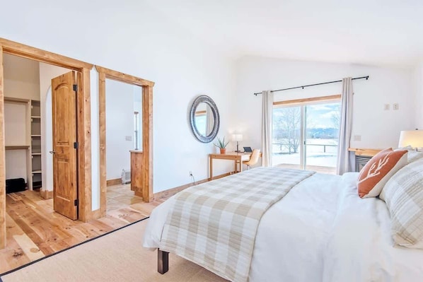 A bedroom with a bed, a desk, a chair, a mirror, and a view of the outdoors.