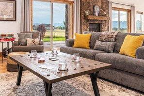 A living room with a stone fireplace, a grey sofa with yellow cushions, a wooden coffee table with glasses and playing cards, and a view of a green landscape through large windows.