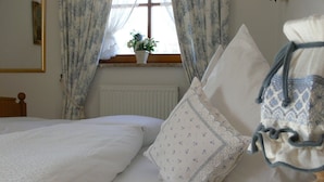 Een slaapkamer met een bed, kussens, een raam met gordijnen, een radiator en een kleine plant op de vensterbank.