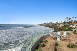 Un patio frente al mar con barandilla de vidrio, mobiliario exterior y vistas al océano y casas costeras.