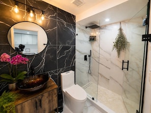 A modern bathroom with a marble wall, a round mirror, a wooden vanity, a white toilet, and a glass-enclosed shower area. - Modern 6BR Home - Jacuzzi, BBQ, Game Room (Miami)