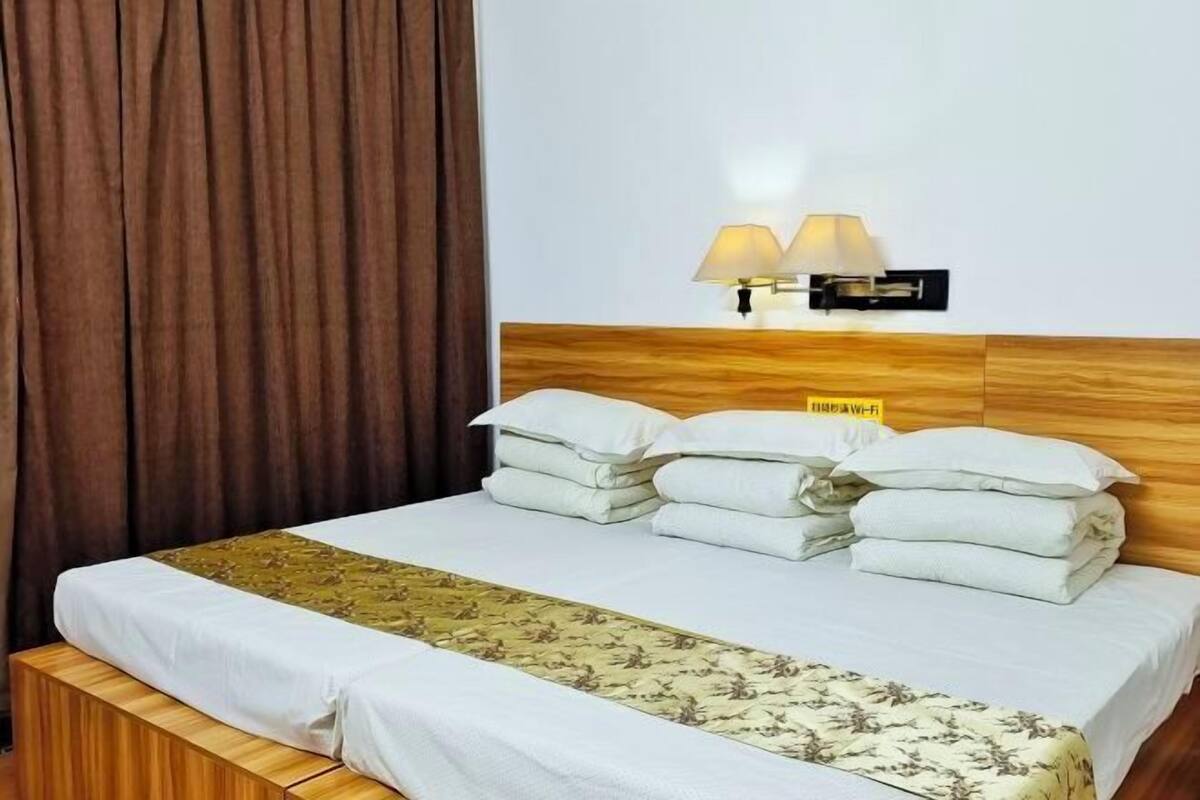 Quarto de hotel com cabeceira de madeira, cama com roupa de cama branca e colcha floral, duas luminárias fixadas na parede e um aviso indicando disponibilidade de Wi-Fi.