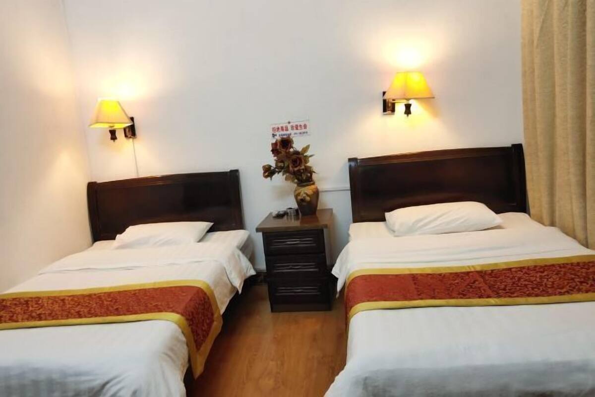 Quarto de hotel com duas camas de solteiro, mesa de cabeceira com luminária e um vaso decorativo na parede.