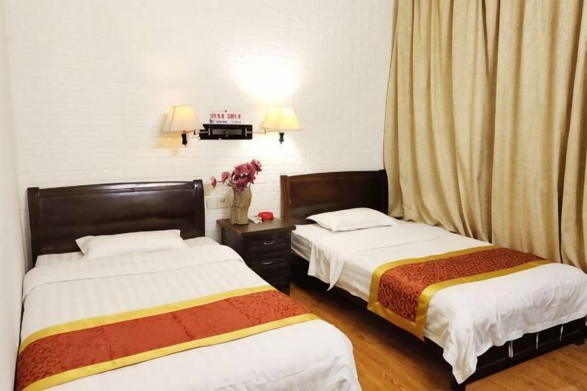 Quarto de hotel com duas camas, uma mesa de cabeceira, um abajur, um relógio na parede e uma cortina.