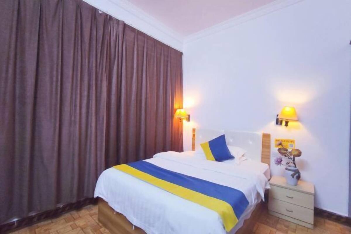 Quarto de hotel com cama, cortinas, abajures, mesa de cabeceira e luminária na parede.