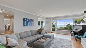 A modern living room with a grey sectional sofa, a piano, and a large window offering a view of the outdoors. - Luxury Water View 6BR Retreat with Gym (Seattle)