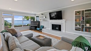 A modern living room with a fireplace, a large TV, and a view of the sea. - Luxury Water View 6BR Retreat with Gym (Seattle)