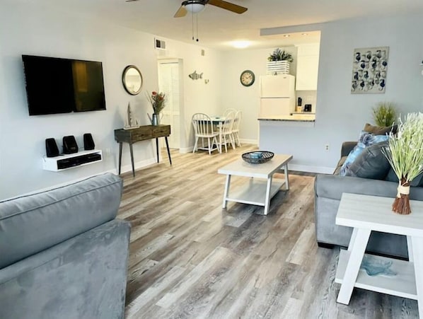 A modern living room with a grey sofa, a white coffee table, and a grey couch. There is a flat-screen TV mounted on the wall, a dining area with a white table and chairs, and a kitchen area with a refrigerator and a sink.