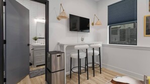 A modern kitchen with a black TV, two bar stools, a trash bin, and a window with blinds. - Cozy Studio in the Crossroads | 69 (Kansas City)
