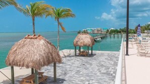 A beachside area with thatched umbrellas, a wooden bench, and a view of the ocean and a pier with buildings in the background. - Ocean Isles Breeze Getaway – Marathon Oi#82 (Marathon)