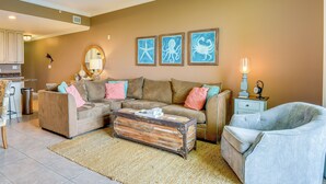 Ein Wohnzimmer mit einem braunen Sofa, einem grauen Sessel, einem Couchtisch und einem Teppich. An der Wand hängen drei gerahmte Bilder.